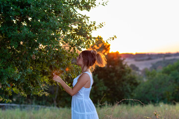 Obraz premium A young girl in profile with long blond tied hair, dressed in white, at sunset, touches the leaves of the magic tree