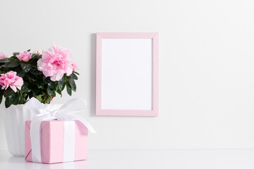 Pink frame mockup on white wall, gift with ribbon, pink flowers in pot decoration. Front view