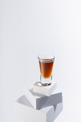 Golden brandy shot glass in hard light with shadow, reflection on high stack of white boxes, alcohol drink in rich elegant minimal geometric style, vertical.