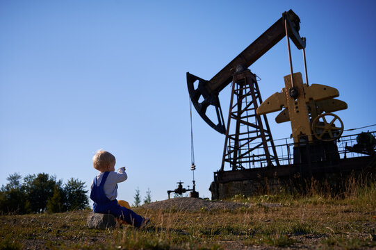 Adorable Child In Work Overalls Sitting On Stone And Pointing At Oil Pump Rocker-machine. Cute Male Kid Oil Worker Looking At Petroleum Pump Jack While Resting Outdoors Under Beautiful Blue Sky.