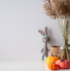 Space for copying on a white background autumn cozy decor crocheted bunny, pumpkins, jute basket, a bouquet of dry ears in a glass vase.