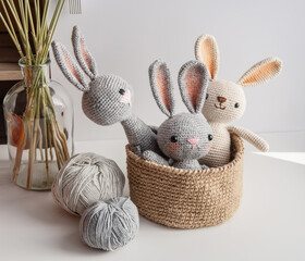 Three handmade bunnies sit in a jute basket on a white table, knitting yarn side by side.