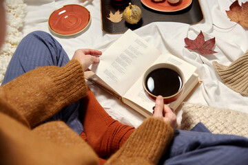 people, season and leisure concept - young woman drinking black coffee and reading book at home in autumn