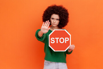 Serious woman with curly hair wearing green casual style sweater showing stop gesture with hand holding red traffic sign of prohibition, blocking. Indoor studio shot isolated on orange background. - obrazy, fototapety, plakaty
