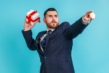 Catch your gift! Young serious businessman wearing dark official style suit, swung and wants to throw off your gift box, pointing finger in distance. Indoor studio shot isolated on blue background.