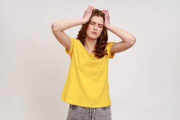 Portrait of sad young woman with curly hair in casual yellow T- shirt standing and holding her painful head with closed eyes, suffering from headache. Indoor studio shot isolated on gray background.