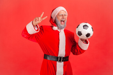 Amazed elderly man with gray beard wearing santa claus costume holding soccer ball in hands, showing v sign to camera, winking, open mouth. Indoor studio shot isolated on red background.