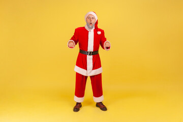 Fototapeta na wymiar Full length portrait of elderly man with gray beard wearing santa claus costume standing with shocking expression and pointing down with both hands. Indoor studio shot isolated on yellow background.