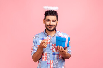 Smiling cute angelic bearded man with nimb over head holding gift box and pointing to camera,...