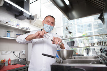 Cheerful chef wearing medical face mask, salting dish in a bowl at his kitchen, copy space