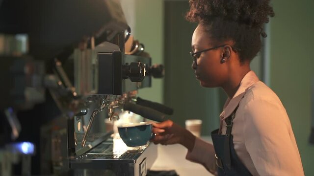 Barista African-American woman with coffeemaker. Spbas Positive black female employee takes cup and puts on equipment board waiting for coffee in cozy cafe