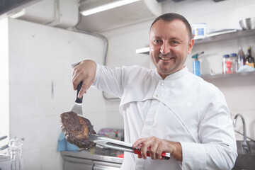 Proud chef showing delicious grilled beef steak to the camera, working at the kitchen