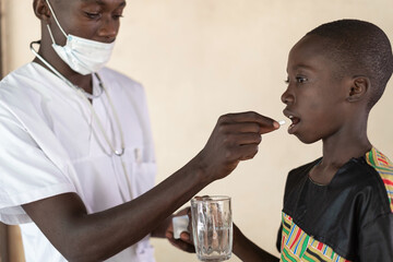 Health professional with protective mask and stethoscope giving a pill to a smal black boy holding...