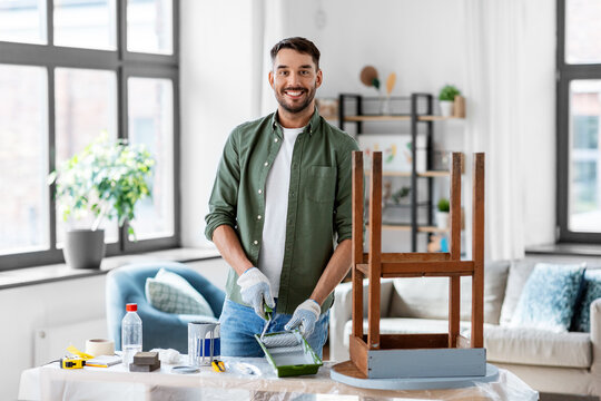 renovation, diy and home improvement concept - happy smiling man in gloves with paint roller painting old wooden table in grey color