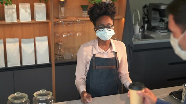 Coffee shop at virus spread prevention. Spbas Afro-American lady in disposable mask gives paper cup and bank terminal to male guest for payment in store