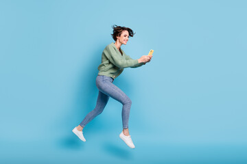Fototapeta na wymiar Photo of pretty hurry young woman wear green cardigan jumping running holding modern device isolated blue color background