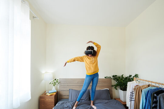 Happy excited teenage girl in virtual reality headset dancing on bed in her room