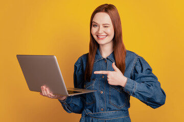 Portrait of cheerful lady hold netbook direct forefinger wink eye promoting gadget on yellow background