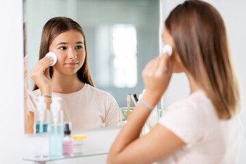 beauty, hygiene and people concept - teenage girl looking in mirror and cleaning face skin with cotton disc at bathroom
