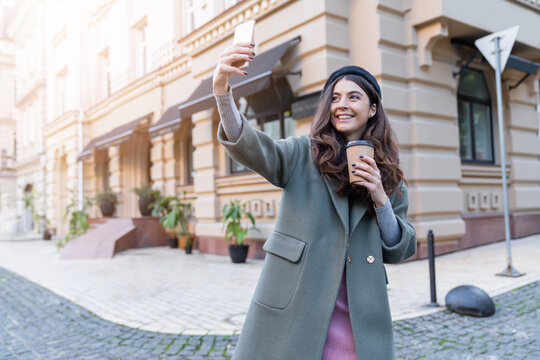 Closeup portrait of shining beautiful brunette in trendy gray trench coat and black beret is walking around autumn city. Pretty young woman is making photo and smiling at her smartphone camera outdoor