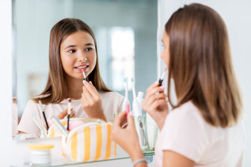 Obraz na płótnie Canvas beauty, make up and cosmetics concept - teenage girl applying lip gloss and looking to mirror at home bathroom