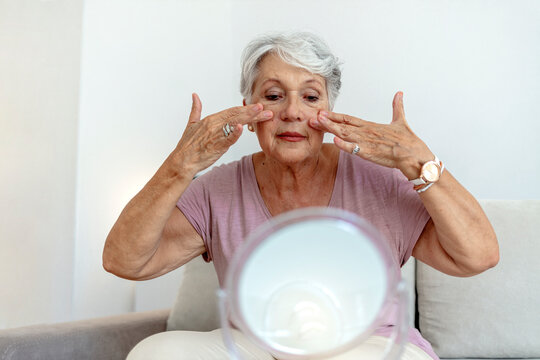 Older Woman Applying Cream To Face And Looking To Mirror At Home. Healthy Skin Care Beauty Routine Treatment Concept. Elegant Old Lady Applying Cream And Looking At Mirror In Modern Home In Sunny Day.