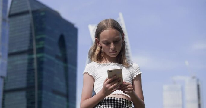 Focused teen girl reading smartphone on glass skyscrapers background in developed city. Beautiful young girl browsing internet content in social networks from mobile phone on urban landscape
