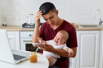 Indoor shot of sad father wearing maroon casual t shirt sitting with baby boy or girl in kitchen, holding smart phone, dialing wife's number, can't calm down daughter or son.