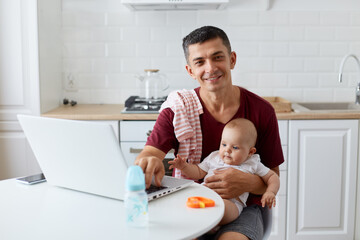 Happy smiling young adult father wearing maroon casual t shirt sitting at table in kitchen near notebook, holding infant in arms, looking at camera with positive expression.