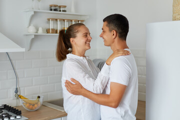 Indoor shot of smiling female hugging with her husband at home in light kitchen, happy couple standing together and looking at each other with love and romantic feelings.