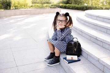 Smiling happy kid girl 5-6 year old wear uniform dress and glasses sitting on stair with books and backpack outdoors. Looking at camera. Back to school concept. Cheerful pupil.
