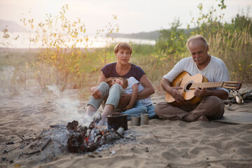 Grandparents and child granddaughter Camping holiday in the summer nature. Camping concept. senior manGrandfather playing guitar and singing song to his Family sitting on the beach near the fire
