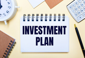 On a light background, a white alarm clock, a calculator, a pen and a notebook with the text INVESTMENT PLAN. Flat lay