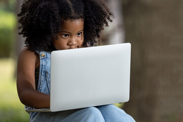 Obraz na płótnie Canvas children girl African American ethnicity black skin sitting on tree base use Laptop computer playing game online with friends via wifi 5G internet signal in the park, social distance concept