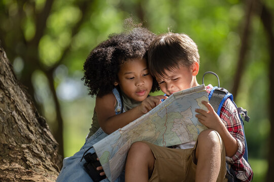 African American ethnicity boy and girl children wearing plaid shirt and backpack sitting at tree base talking and looking down on the map travel on hand in the park 
