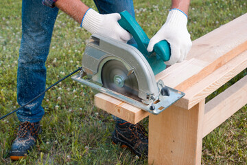 A man builder with an electric hand-held circular saw saws a wooden board on the lawn. DIY in the backyard.