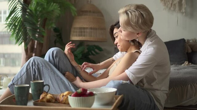 Lesbian couple after breakfast in bedroom. Spbd Young Asian woman with blonde wife spends time hugging on floor near comfortable bed in lazy morning side view