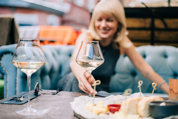 Happy and laughing adult mature woman sitting in bar outdoors with wine glasses and blurry restaurant background scene, drinking white wine and eating cheese. Summer sunny day on patio.