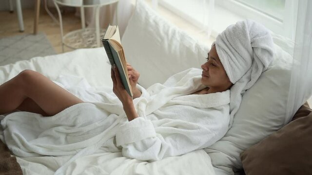 Beauty and body care at home. Spbd Happy African-American lady in bathrobe with towel turban on head reads book lying on comfortable bed near window