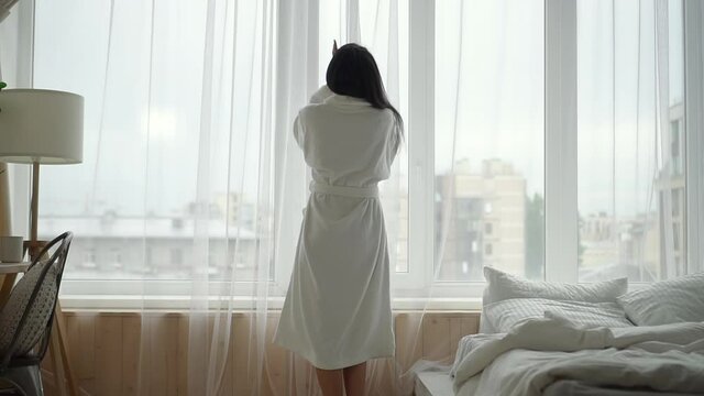 Beauty healthcare luxury spa resort. Spbd African-American woman wearing long terry bathrobe opens window curtains in hotel room backside view
