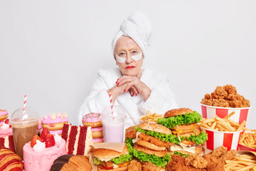 Serious old woman has cheat meal day at home keeps hands under chin affords eating tasty burgers doughnuts and cakes applies beauty patches under eyes to reduce wrinkles wears soft white bathrobe