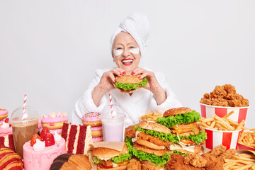 Positive old woman with red lips enjoys eating tasty hamburger addicted to junk food doesnt keep to diet applies beauty patches under eyes poses at table feels hungry. Cheat meal consumption