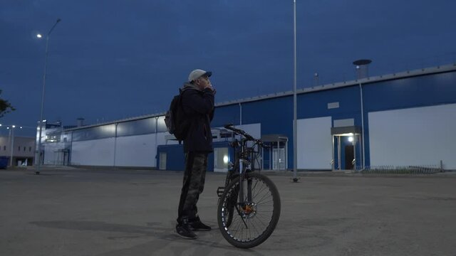 A Man with a Bicycle Stands Among the Night City and Plays the Vargan. Cyclist Illuminated by Night Lanterns. Ethnic Connection with Timeliness. Musician Artist Relaxing Outdoors