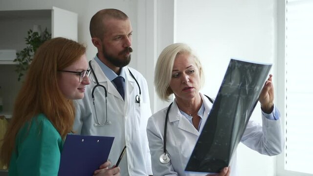 Doctor and colleagues in hospital. Spbd Middle aged lady traumatologist shows x-ray picture of patient to younger general practitioners in clinic office