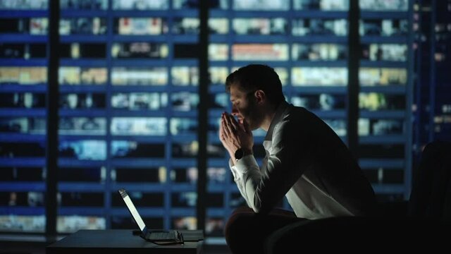 Tired young man working on a laptop late night in the office. Sleepy Businessman sitting at desk in dark office. Tired and stressed businessman in glasses works on a laptop of the night city office