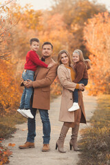 Happy family hugging on a walk in the fall park. Portrait of a caucasian mother and father holding their children in beautiful outfits on a sunny autumn day in forest. Family lifestyle concept.