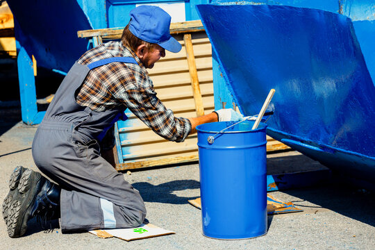 An old man in a work jumpsuit paints a garbage can or container on the street with a paint brush. Part-time work for a pensioner. Toil.