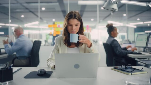 Young Happy Businesswoman Using Computer in Modern Office with Colleagues. Stylish Beautiful Manager Smiling, Working on Financial and Marketing Projects. Drinking Tea or Coffee from a Mug.