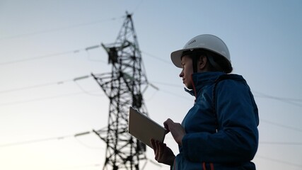 A woman energy worker in a white helmet works checks the power line using a computer on a tablet,...