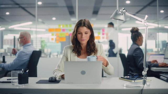 Young Happy Businesswoman Using Computer in Modern Office with Colleagues. Stylish Beautiful Manager Smiling, Working on Financial and Marketing Projects. Drinking Tea or Coffee from a Mug.